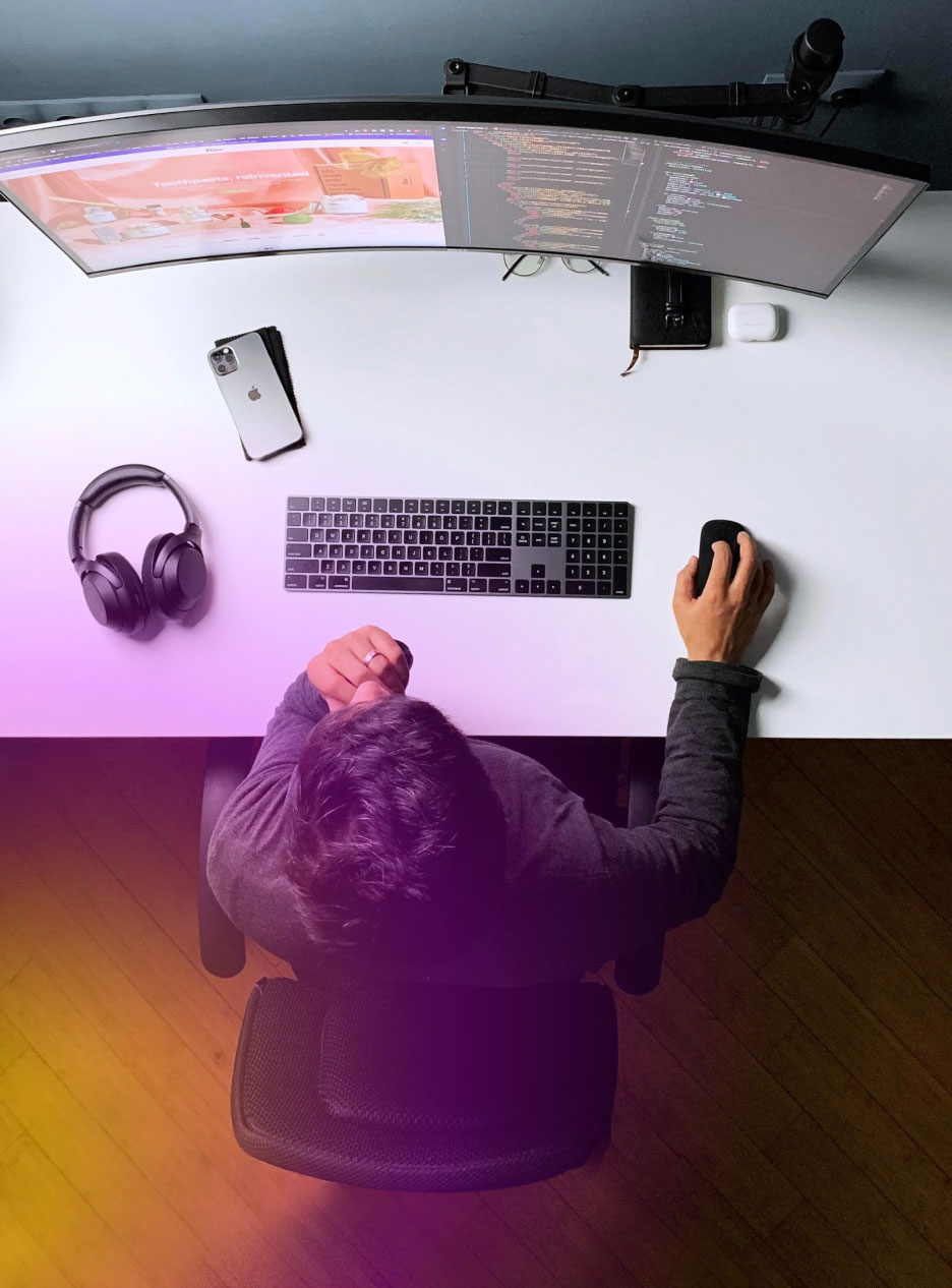 info-14 Büro von oben. Mann am Schreibtisch mit Tastatur, Maus, Handy, Kopfhörern und einem großen geschwundenen Bildschirm.