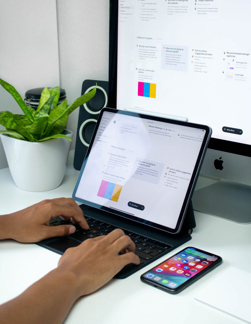 info-6 Arbeitsplatz mit Bildschirm, Laptop, Handy und Pflanze.
